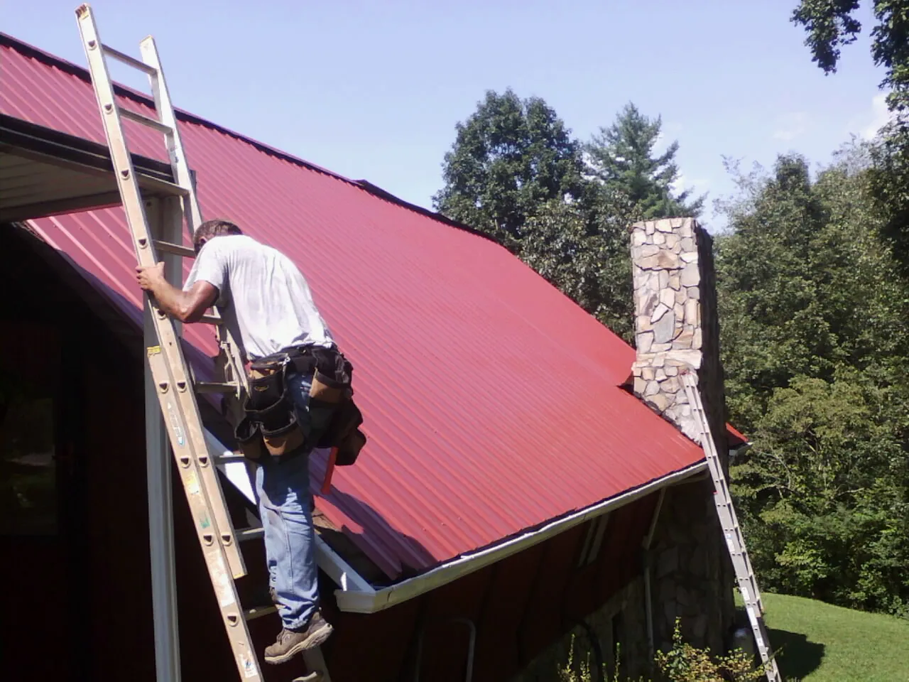 Roofer on a ladder beside a red metal roof and stone chimney — Soffit & Fascia Repair by Roofing Brothers in Poplar Branch